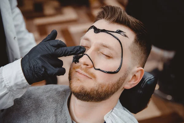 Spa and skin care for men. Barber professional applies black charcoal mask to client man — 스톡 사진