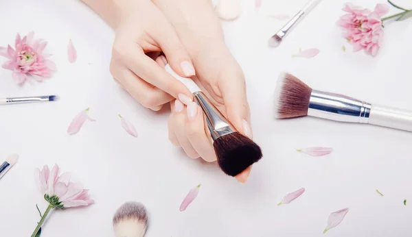 Las manos de las niñas sostienen pinceles de maquillaje en el fondo de flores de color rosa, concepto de salón de belleza —  Fotos de Stock