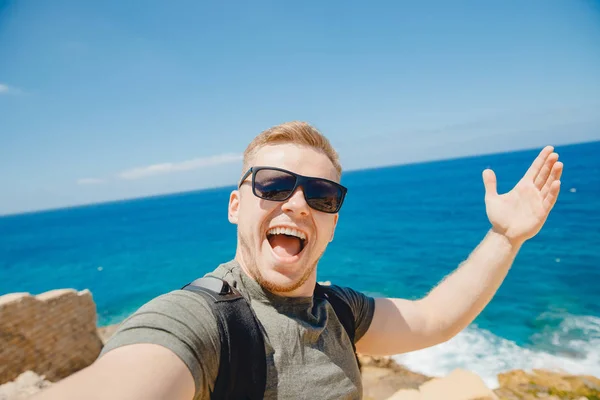 Szczęśliwy podróżnik robi sobie selfie w okularach przeciwsłonecznych na tle stłuczonego morza z plecakiem. Koncepcja podróży — Zdjęcie stockowe