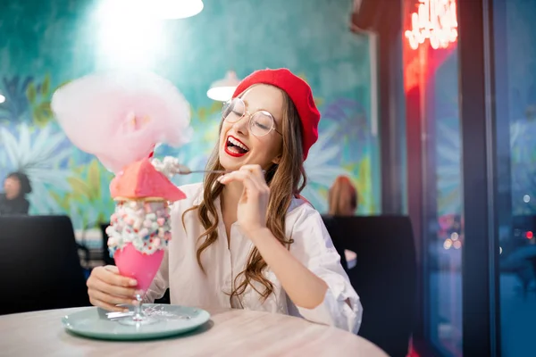 Junge Frau mit roter Baskenmütze und Gläsern hat süßes Dessert mit Zuckerwatte, Milchshake, Knödel und rosa Kaugummikuchen im französischen Café — Stockfoto