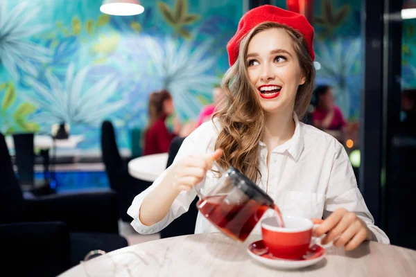 Image romantique de la France. Jeune fille au béret rouge aux cheveux ondulés sourit et verse le thé pour le petit déjeuner dans une tasse dans un café — Photo