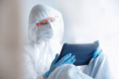 Koronavirüs karantinası sırasında internet üzerinden iletişim. Koruyucu giysili ve tıbbi maskeli kız tablet web kameralı sohbet tutuyor