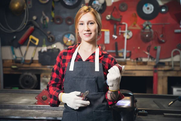 Hermosa mujer joven ingeniero artesano se encuentra en delantal en el fondo de las herramientas para la elaboración. Concepto de pequeña empresa en garaje sala industrial — Foto de Stock