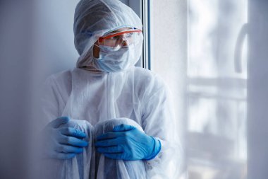 Yalnızlık karantinası, tıbbi maskeli genç bir kadın pencereden kendini izole ediyor. Coronavirüs salgını konsepti