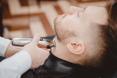 Berber dükkanında saç örgüsü yapan adama sakal kırpıyor. Elektrikli tıraş makinesi, renkli kahverengi.