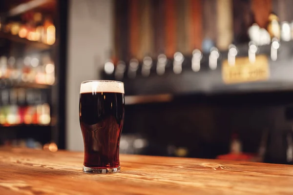 Bicchiere di birra scura artigianale con schiuma sul bancone del bar in legno, pub sfondo sfocato — Foto Stock