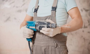 Portrait man builder repairer in protective glasses, hard hat, overalls showing having equipment in arm on grey background home brick