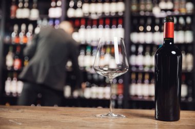 Şişe kırmızı şarap ve cam kadeh ahşap masada, arka planda şarap garsonu restoranın rafından içmeyi seçiyor.