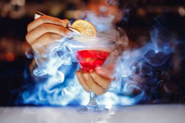 Barmen kırmızı alkollü kokteyl hazırlıyor martini bardağında dumanla, portakalla süslenmiş.