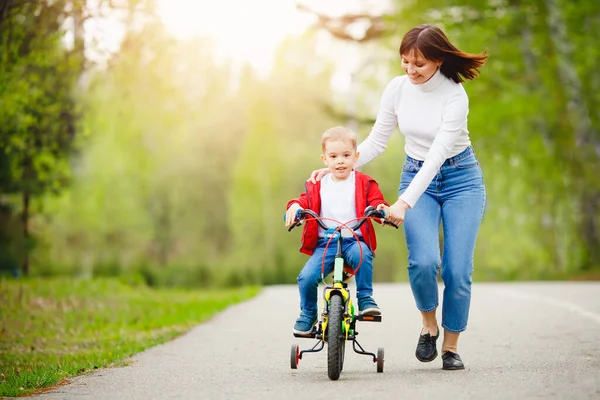 A mãe ensina o filho a andar de bicicleta no parque, manter o equilíbrio, se divertir com a família. Conceito de dia mãe — Fotografia de Stock