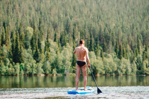 Hombre remo remo en sup board azul lago agua paddleboard fondo de bosque — Foto de Stock