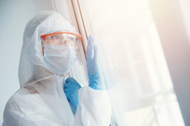 Yalnızlık karantinası, tıbbi maskeli genç bir kadın pencereden kendini izole ediyor. Coronavirüs salgını konsepti