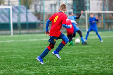 Kırmızı ve mavi spor giyimli çocuklar sahada futbol oynuyor, top sürüyor. Yeşil çimlerde top oynayan genç futbolcular. Eğitim, futbol, çocuk konsepti için aktif yaşam tarzı