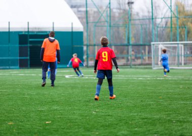 Kırmızı ve mavi spor giyimli çocuklar sahada futbol oynuyor, top sürüyor. Yeşil çimlerde top oynayan genç futbolcular. Eğitim, futbol, çocuk konsepti için aktif yaşam tarzı