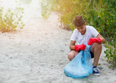 Eldivenli beyaz tişörtlü çocuk sahilde ki mavi pakette çöp ve plastik şişeleri topluyor. Genç gönüllü. Çevre koruma, çevre kavramını korumak. Eldivenle ellerinizi kapatın