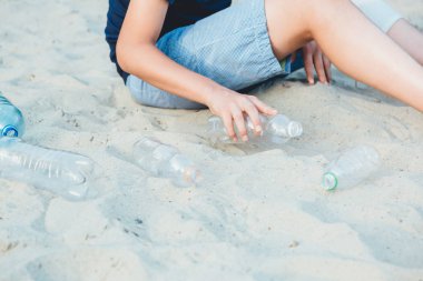 Mavi tişörtlü gönüllü çocuk parkta kirli plastik şişeleri topluyor. Sarı eldivenli eller çöp toplar, torbadaki çöpleri toplar. Plastikten doğayı temizleyen çevreci eylemci