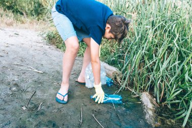 Mavi tişörtlü gönüllü çocuk parkta kirli plastik şişeleri topluyor. Sarı eldivenli eller çöp toplar, torbadaki çöpleri toplar. Plastikten doğayı temizleyen çevreci eylemci