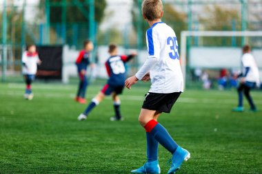 Beyaz ve mavi spor giyen çocuklar sahada futbol oynar, top top akıtır. Yeşil çimüzerinde topu ile genç futbolcular. Eğitim, futbol, çocuklar için aktif yaşam tarzı kavramı