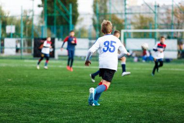 Beyaz ve mavi spor giyen çocuklar sahada futbol oynar, top top akıtır. Yeşil çimüzerinde topu ile genç futbolcular. Eğitim, futbol, çocuklar için aktif yaşam tarzı kavramı