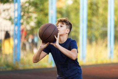 Mavi tişörtlü şirin çocuk şehir parkında basketbol oynuyor. Aktif gençler turuncu topla açık havada oyun oynuyorlar. Hobi, aktif yaşam tarzı, çocuklar için spor.