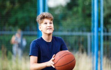 Mavi tişörtlü şirin çocuk şehir parkında basketbol oynuyor. Aktif gençler turuncu topla açık havada oyun oynuyorlar. Hobi, aktif yaşam tarzı, çocuklar için spor.