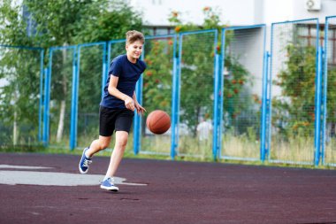 Mavi tişörtlü şirin çocuk şehir parkında basketbol oynuyor. Aktif gençler turuncu topla açık havada oyun oynuyorlar. Hobi, aktif yaşam tarzı, çocuklar için spor.