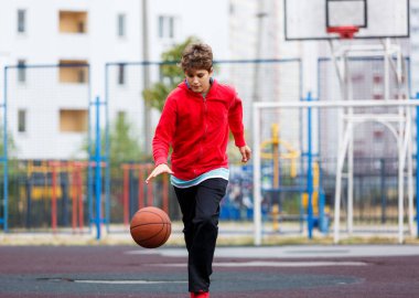 Kırmızı tişörtlü şirin çocuk şehir parkında basketbol oynuyor. Aktif gençler turuncu topla açık havada oyun oynuyorlar. Hobi, aktif yaşam tarzı, çocuklar için spor.