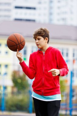 Kırmızı tişörtlü şirin çocuk şehir parkında basketbol oynuyor. Aktif gençler turuncu topla açık havada oyun oynuyorlar. Hobi, aktif yaşam tarzı, çocuklar için spor.