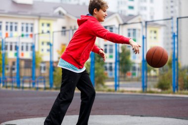 Sevimli gülümseyen çocuk şehir parkında basketbol oynuyor. Aktif gençler turuncu topla açık havada oyun oynuyorlar. Hobi, aktif yaşam tarzı, çocuklar için spor, gençler