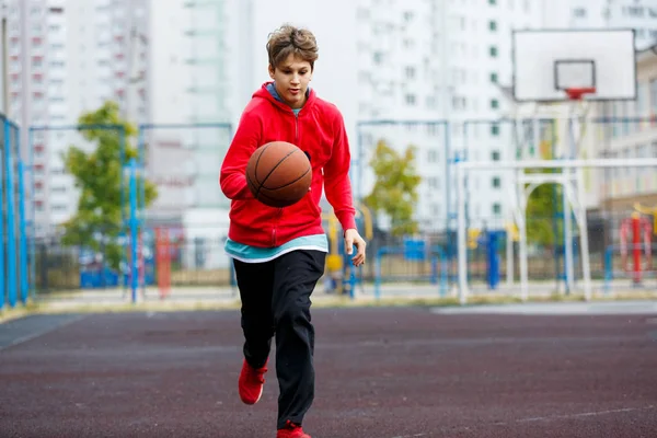 Cute smiling boy plays basketball on city playground. Active teen enjoying outdoor game with orange ball. Hobby, active lifestyle, sport for kids, teenagers