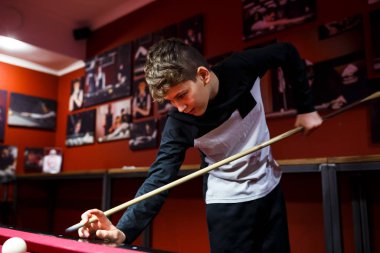 Boy plays billiard or pool in club. Young Kid learns to play snooker. Boy with billiard cue strikes the ball on table. Active Leisure, sport, hobby concept.
