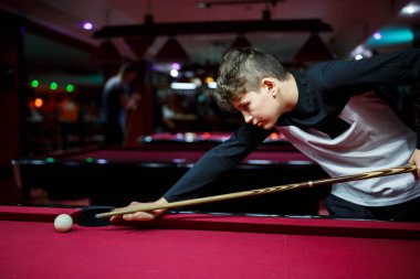 Boy plays billiard or pool in club. Young Kid learns to play snooker. Boy with billiard cue strikes the ball on table. Active Leisure, sport, hobby concept.