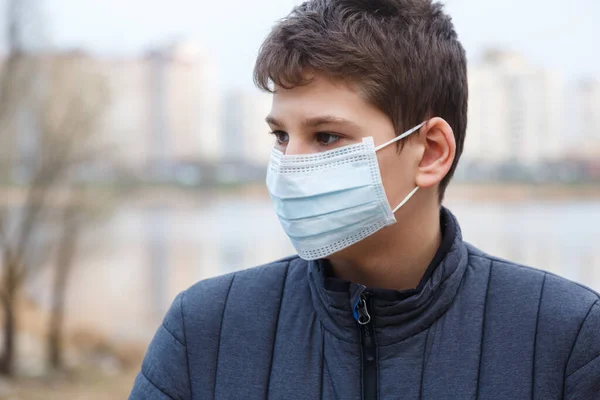 Young cute caucasian boy wearing protective mask against the corona virus on the street. Teenager in surgical face mask to prevent from virus Covid 19 in the city. Close up portrait.