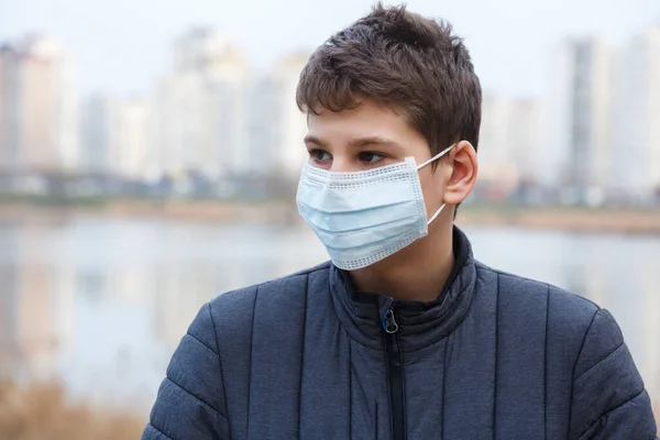 Young cute caucasian boy wearing protective mask against the corona virus on the street. Teenager in surgical  face mask to prevent from virus Covid 19 in the city.  Close up portrait