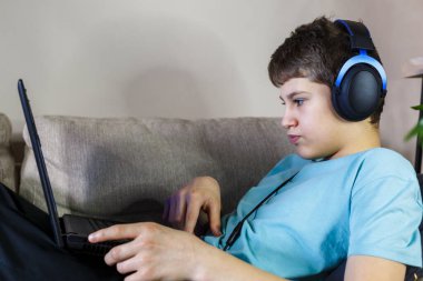 Oturma odasındaki kanepede oturan yeşil tişörtlü yakışıklı çocuk dizüstü bilgisayarıyla ders çalışıyor. Evde eğitim, kişisel eğitim, çocukların uzaktan öğrenme.