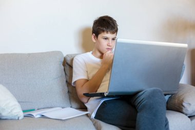 Gözlüklü ve beyaz tişörtlü yakışıklı çocuk oturma odasındaki kanepede dizüstü bilgisayarla oturuyor ve ders çalışıyor. Evde eğitim, kişisel eğitim, çocukların uzaktan öğrenme.