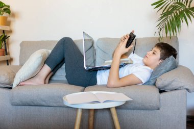 Gözlüklü, beyaz tişörtlü, dizüstü bilgisayarı ve telefonuyla oturma odasındaki kanepede yatan tatlı çocuk. Evde eğitim, kişisel eğitim, çocukların uzaktan öğrenme.