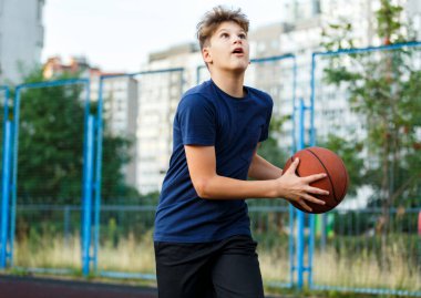 Mavi tişörtlü sevimli çocuk şehir parkında basketbol oynuyor. Aktif gençler turuncu topla açık havada oyun oynuyorlar. Hobi, aktif yaşam tarzı, çocuklar için spor.