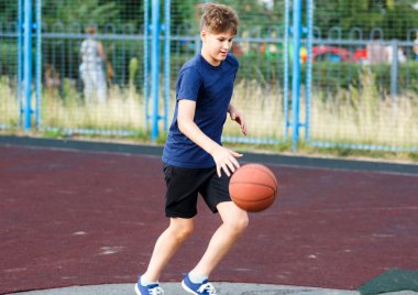 Mavi tişörtlü şirin çocuk şehir parkında basketbol oynuyor. Aktif gençler turuncu topla açık havada oyun oynuyorlar. Hobi, aktif yaşam tarzı, çocuklar için spor, gençler.