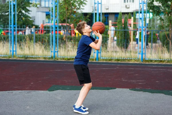 Mavi tişörtlü sevimli çocuk şehir parkında basketbol oynuyor. Aktif gençler turuncu topla açık havada oyun oynuyorlar. Hobi, aktif yaşam tarzı, çocuklar için spor.