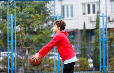 Kırmızı ceketli sevimli gülümseyen çocuk şehir parkında basketbol oynuyor. Aktif gençler turuncu topla açık havada oyun oynuyorlar. Hobi, aktif yaşam tarzı, çocuklar için spor.