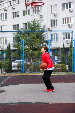 Kırmızı ceketli sevimli gülümseyen çocuk şehir parkında basketbol oynuyor. Aktif gençler turuncu topla açık havada oyun oynuyorlar. Hobi, aktif yaşam tarzı, çocuklar için spor.