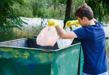 Eldivenli mavi tişörtlü çocuk çöpleri ve plastik şişeleri plajda paketlere doldurup konteynıra atıyor. Genç gönüllü. Çevre koruma, çevre konseptini kaydet
