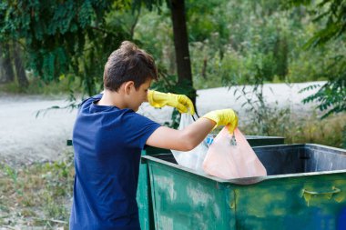 Eldivenli mavi tişörtlü çocuk çöpleri ve plastik şişeleri plajda paketlere doldurup konteynıra atıyor. Genç gönüllü. Çevre koruma, çevre konseptini kaydet