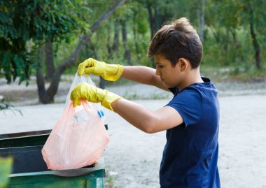 Eldivenli beyaz tişörtlü çocuk plajda çöp ve plastik şişeler topluyor. Genç gönüllü. Çevre koruma, çevre konseptini kaydet