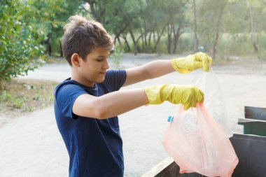 Eldivenli mavi tişörtlü çocuk çöpleri ve plastik şişeleri plajda paketlere doldurup konteynıra atıyor. Genç gönüllü. Çevre koruma, çevre konseptini kaydet