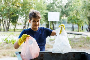 Eldivenli mavi tişörtlü çocuk çöpleri ve plastik şişeleri plajda paketlere doldurup konteynıra atıyor. Genç gönüllü. Çevre koruma, çevre konseptini kaydet