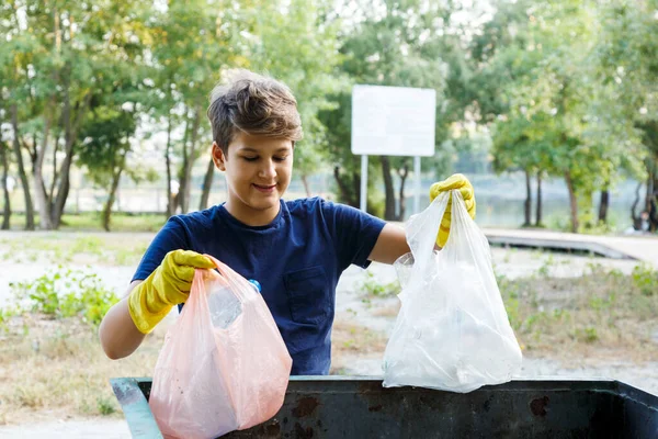 Eldivenli mavi tişörtlü çocuk çöpleri ve plastik şişeleri plajda paketlere doldurup konteynıra atıyor. Genç gönüllü. Çevre koruma, çevre konseptini kaydet