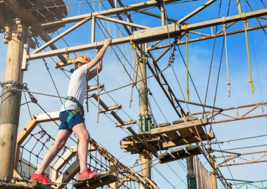 Orman macera parkında bir çocuk. Turuncu kasklı ve beyaz tişörtlü çocuk yüksek ip izine tırmanıyor. Çeviklik becerileri ve çocuklar için açık hava eğlence merkezine tırmanma. Genç çocuk ağaçların tepesinde oynuyor.
