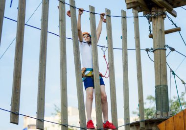 Orman macera parkında bir çocuk. Turuncu kasklı ve beyaz tişörtlü çocuk yüksek ip izine tırmanıyor. Çeviklik becerileri ve çocuklar için açık hava eğlence merkezine tırmanma. Genç çocuk ağaçların tepesinde oynuyor.